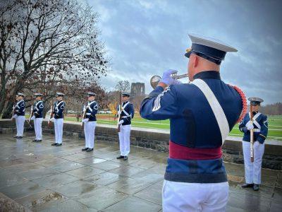 A cadet bugler plays Taps during a wreath-laying ceremony at the Pylons while cadets in dress uniform stand at attention.