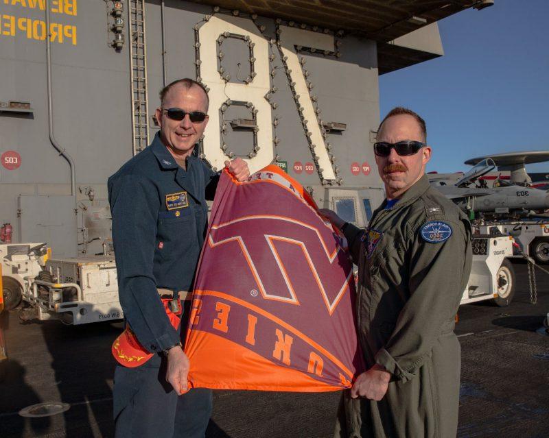Dartez and Von St. Paul stand with a small Virginia Tech flag on the deck of a Navy ship. They are both smiling and wearing military coveralls and sunglasses. 